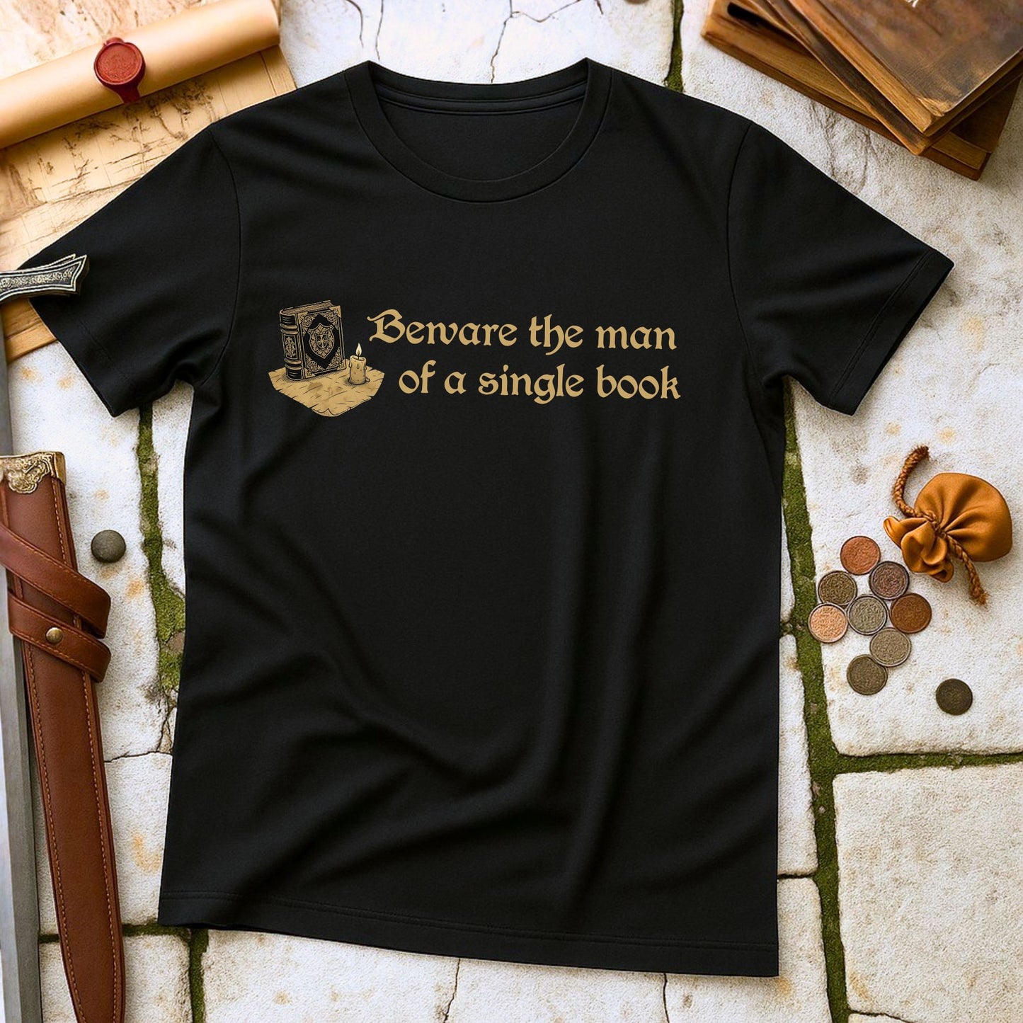 Black t-shirt with gold text 'Beware the man of a single book' on a stone surface with books and coins.