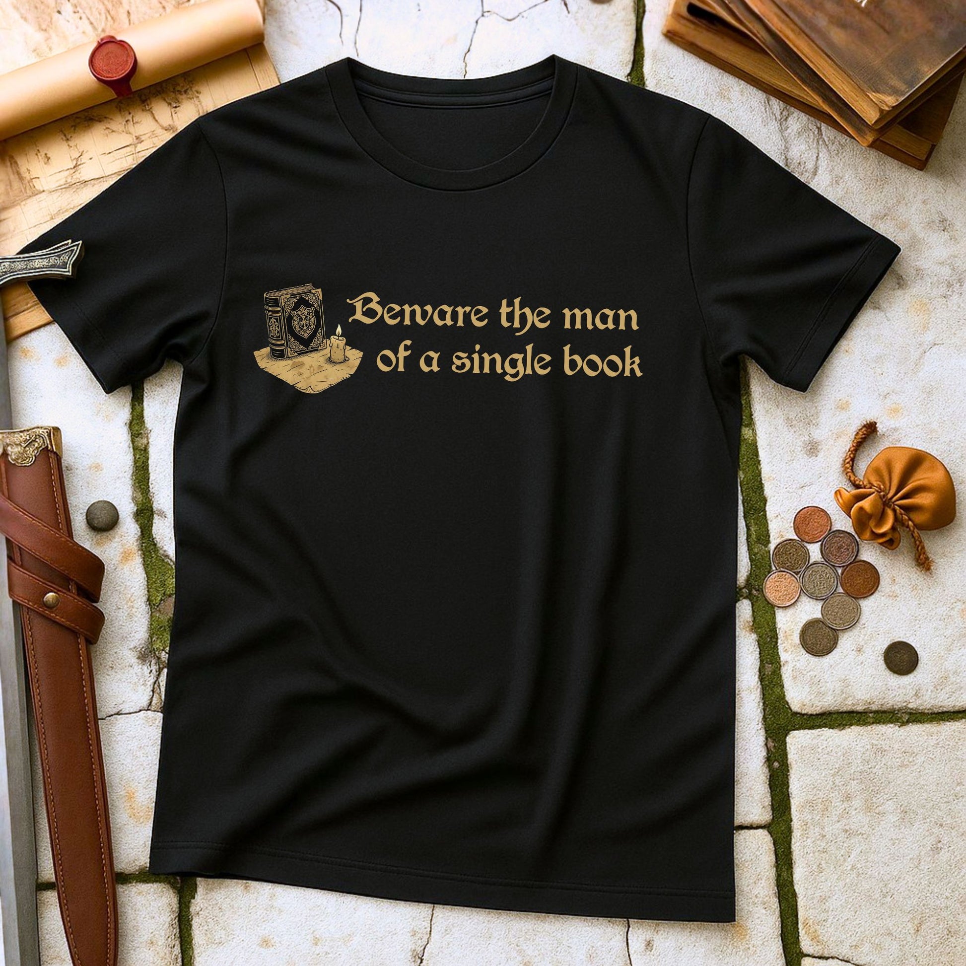 Black t-shirt with gold text 'Beware the man of a single book' on a stone surface with books and coins.